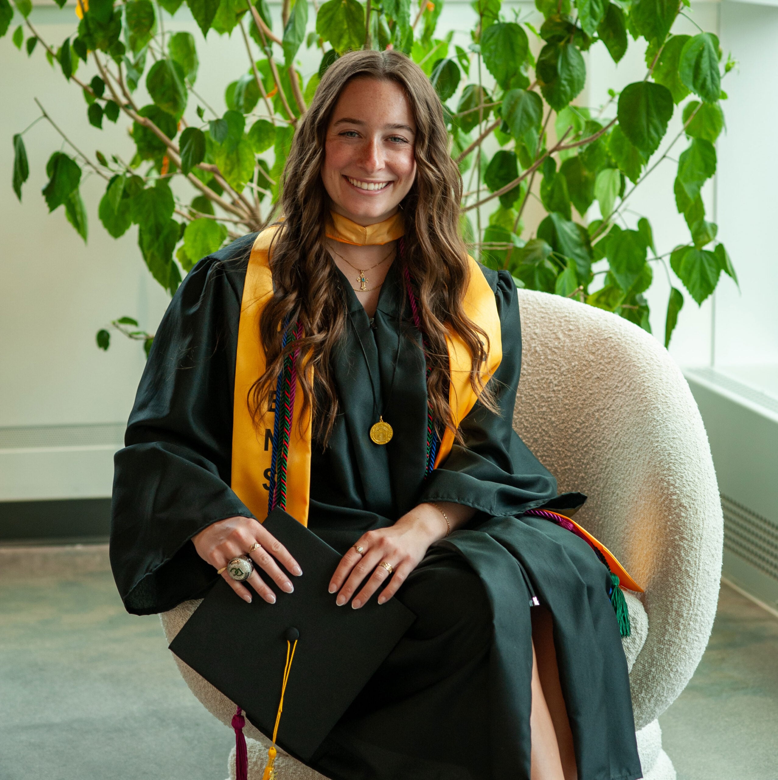 Victoria P. in a graduation gown and sash sits in a chair, holding a diploma, with a plant in the background.