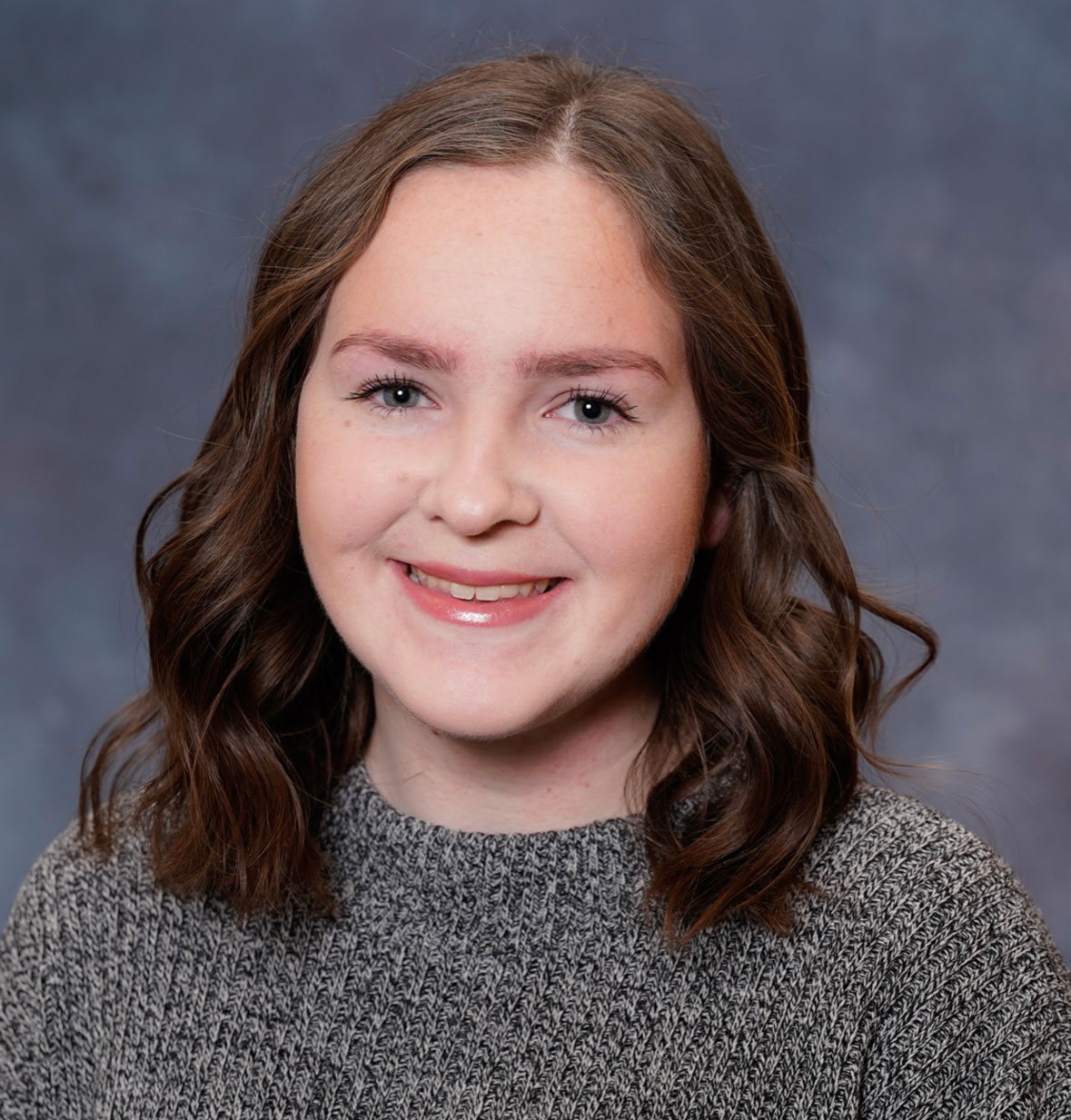 Sophie L. with wavy hair smiles in a close-up portrait against a blurred gray background.