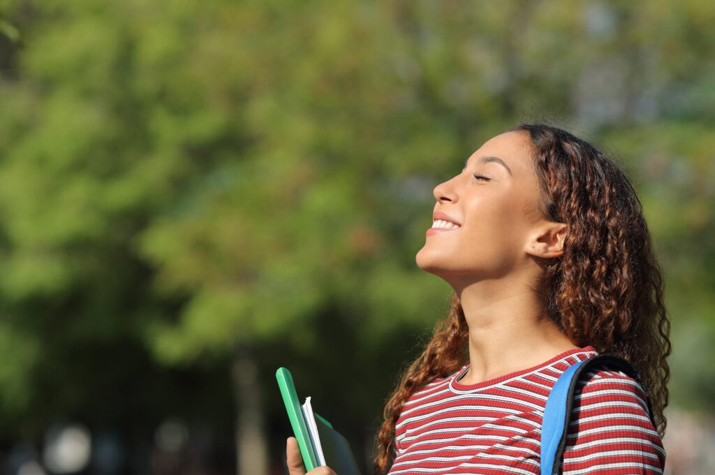 A person stands outdoors with eyes closed, smiling and holding notebooks, surrounded by greenery.
