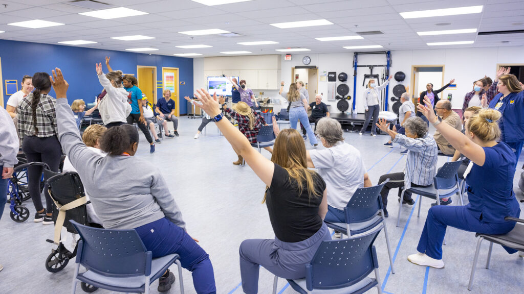Students and community members raise their arms during a seated group exercise class in a UM-Flint classroom.