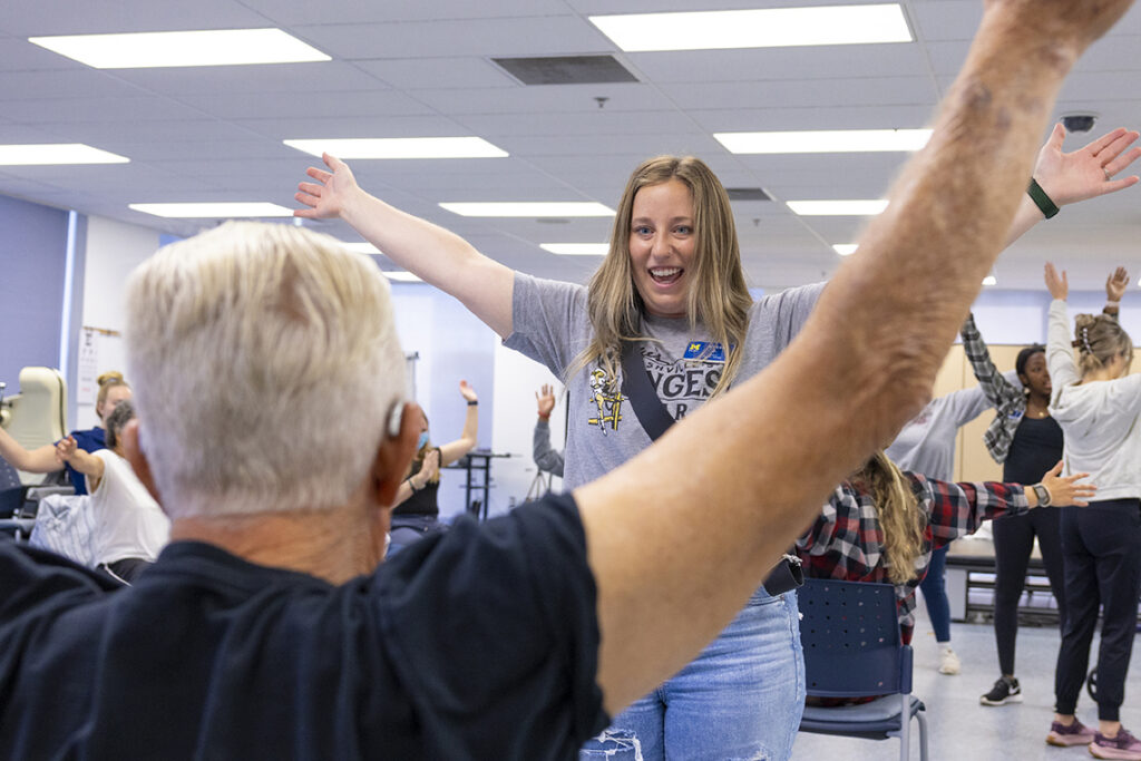 A person with outstretched arms leads a group in an exercise class, with participants following along in a bright room.