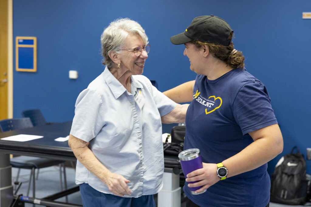 Two people are smiling and embracing in a room with blue walls, one holding a drink and wearing a cap.
