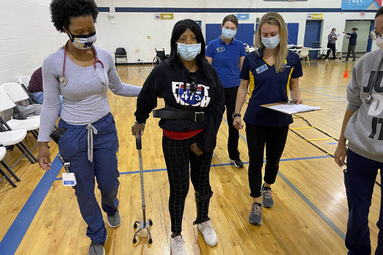 A person using crutches is assisted by two individuals in a gym setting, while another person observes with a clipboard.