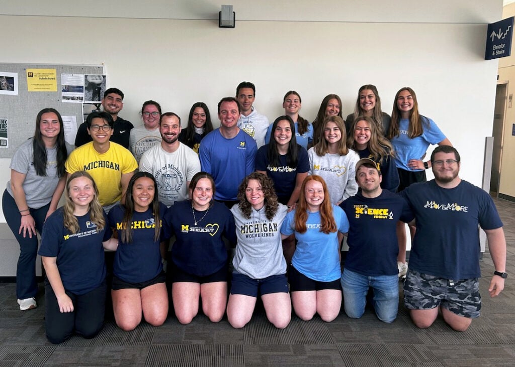 Twenty-two UM-Flint students and faculty members pose together indoors, smiling and wearing maize-and-blue apparel.