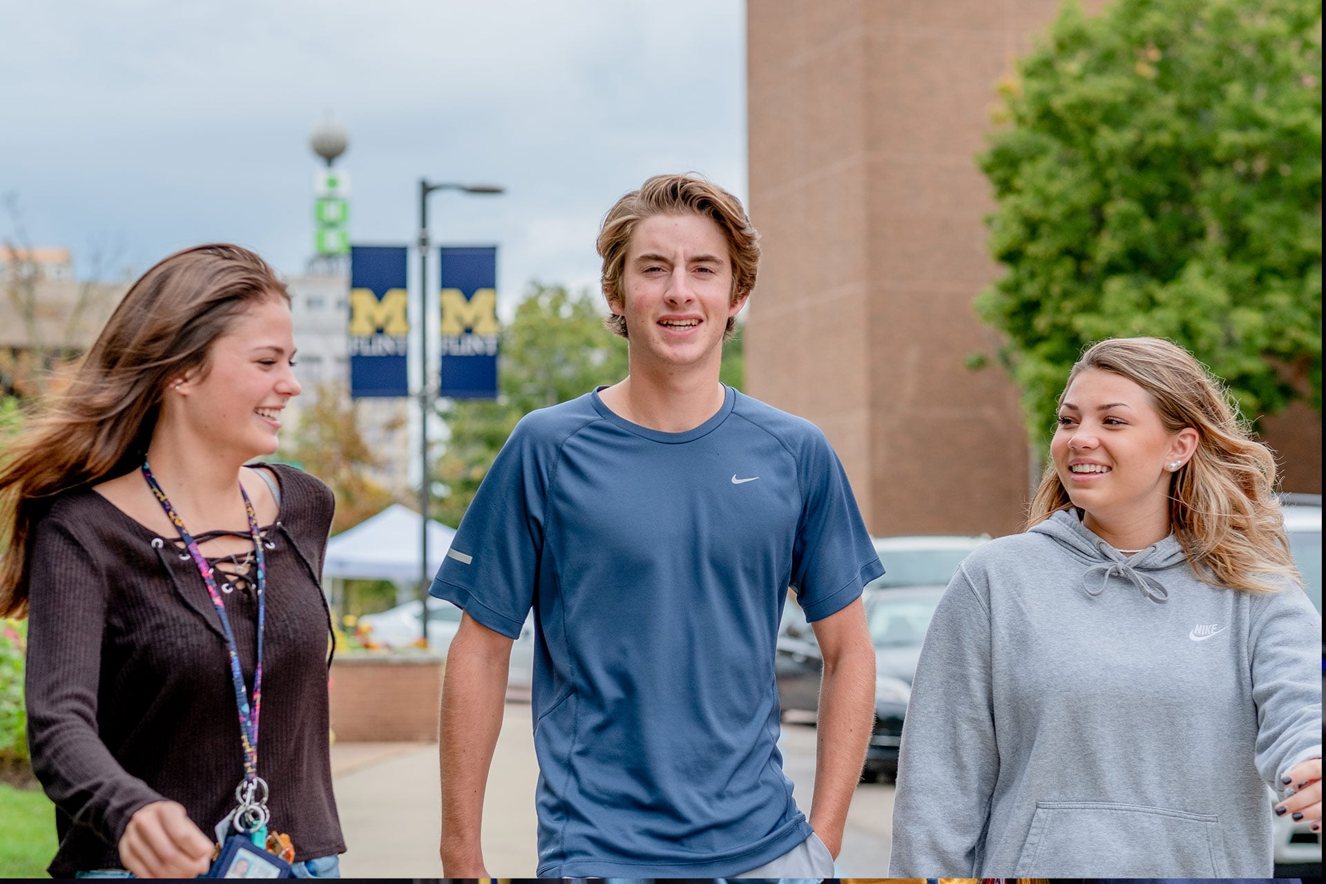 Three people are walking together outdoors, with a university banner visible in the background.