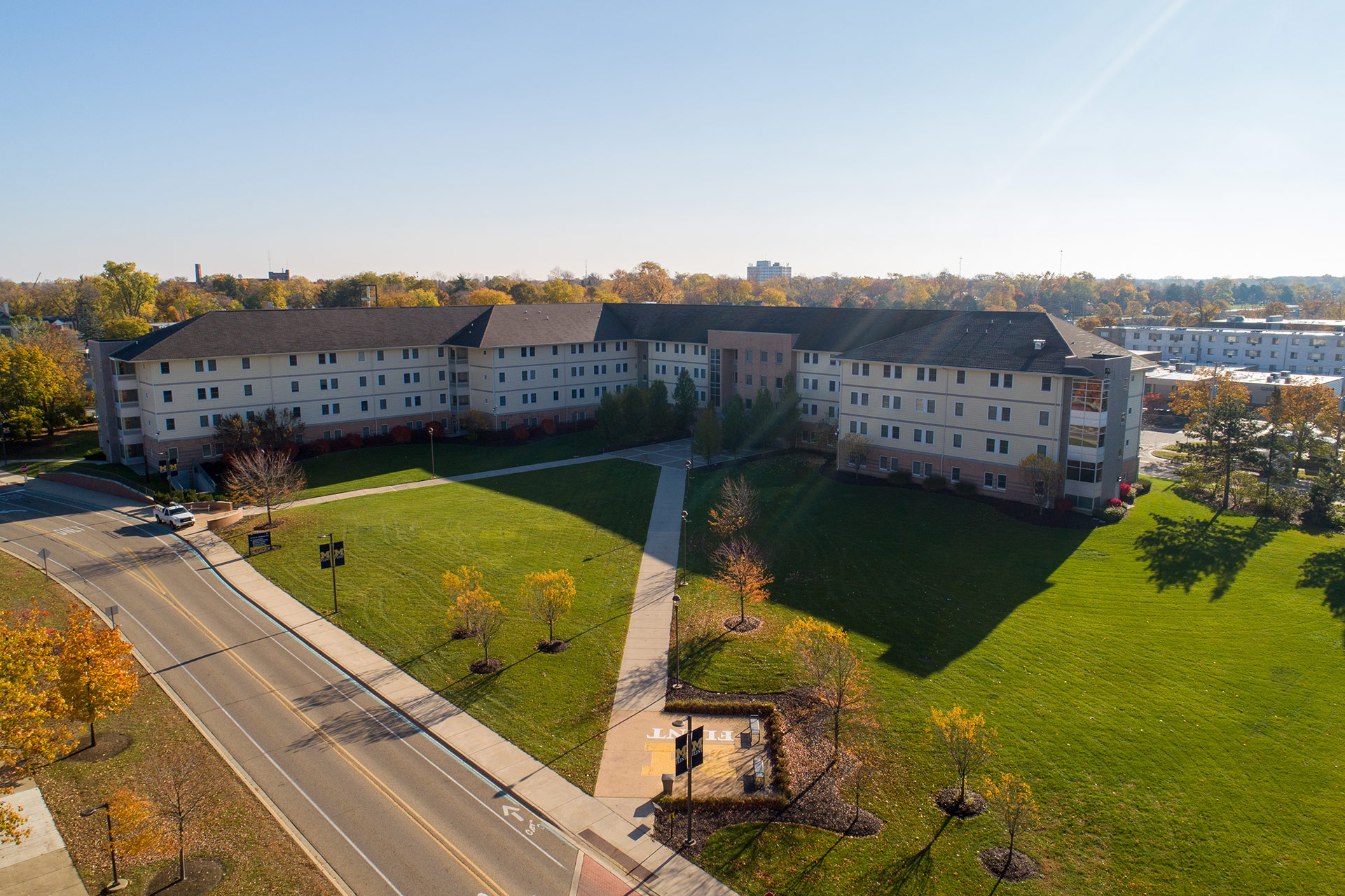 Aerial view of a multi-story residential building surrounded by green grass and trees, with a road and pathway in front.