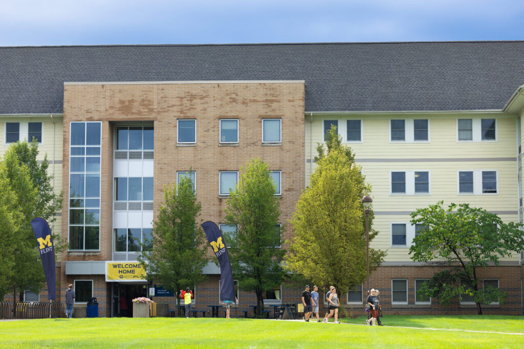 University of Michigan-Flint residence hall with people walking on the grass and a "Welcome Home" banner at the entrance.