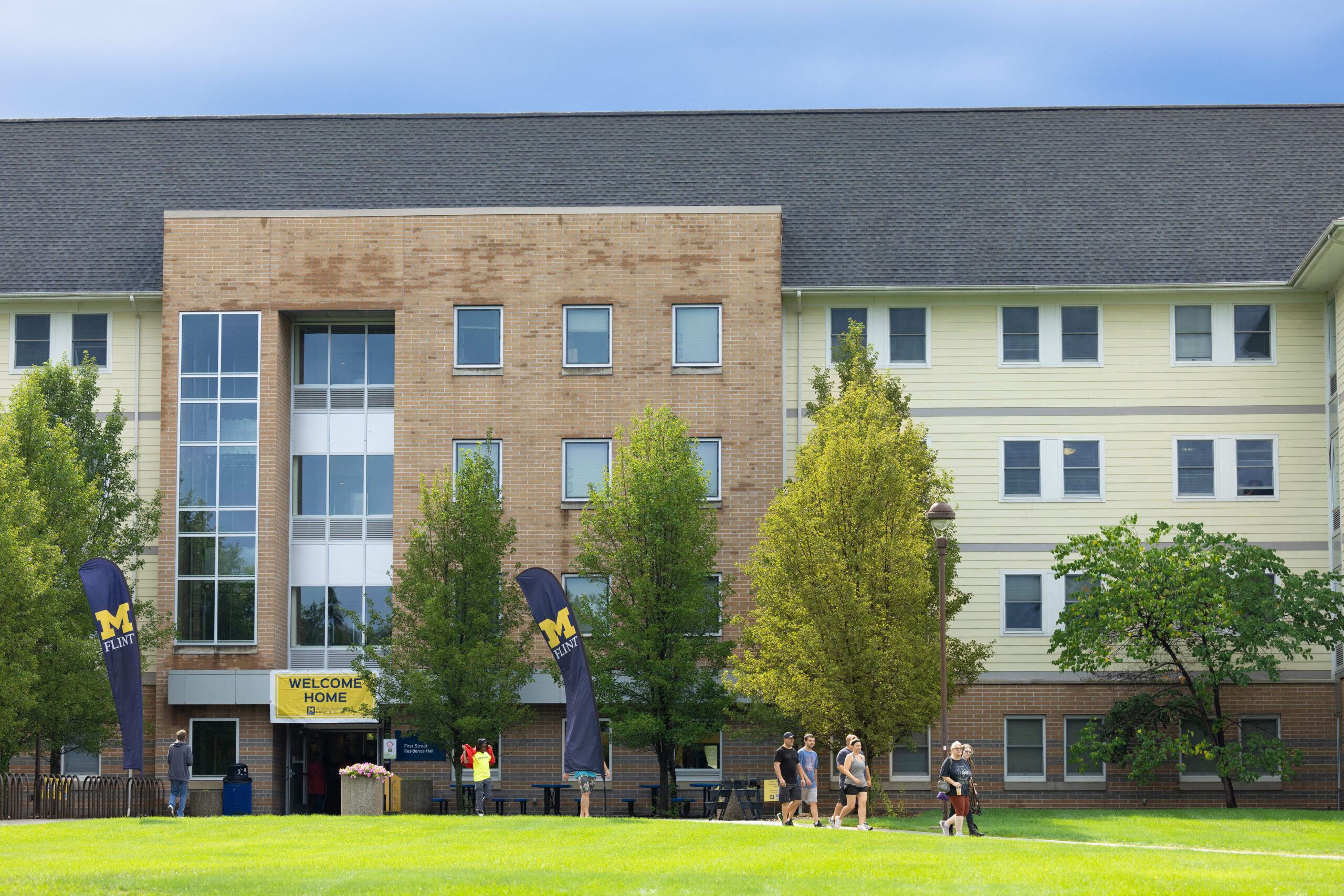 University of Michigan-Flint residence hall with people walking on the grass and a "Welcome Home" banner at the entrance.