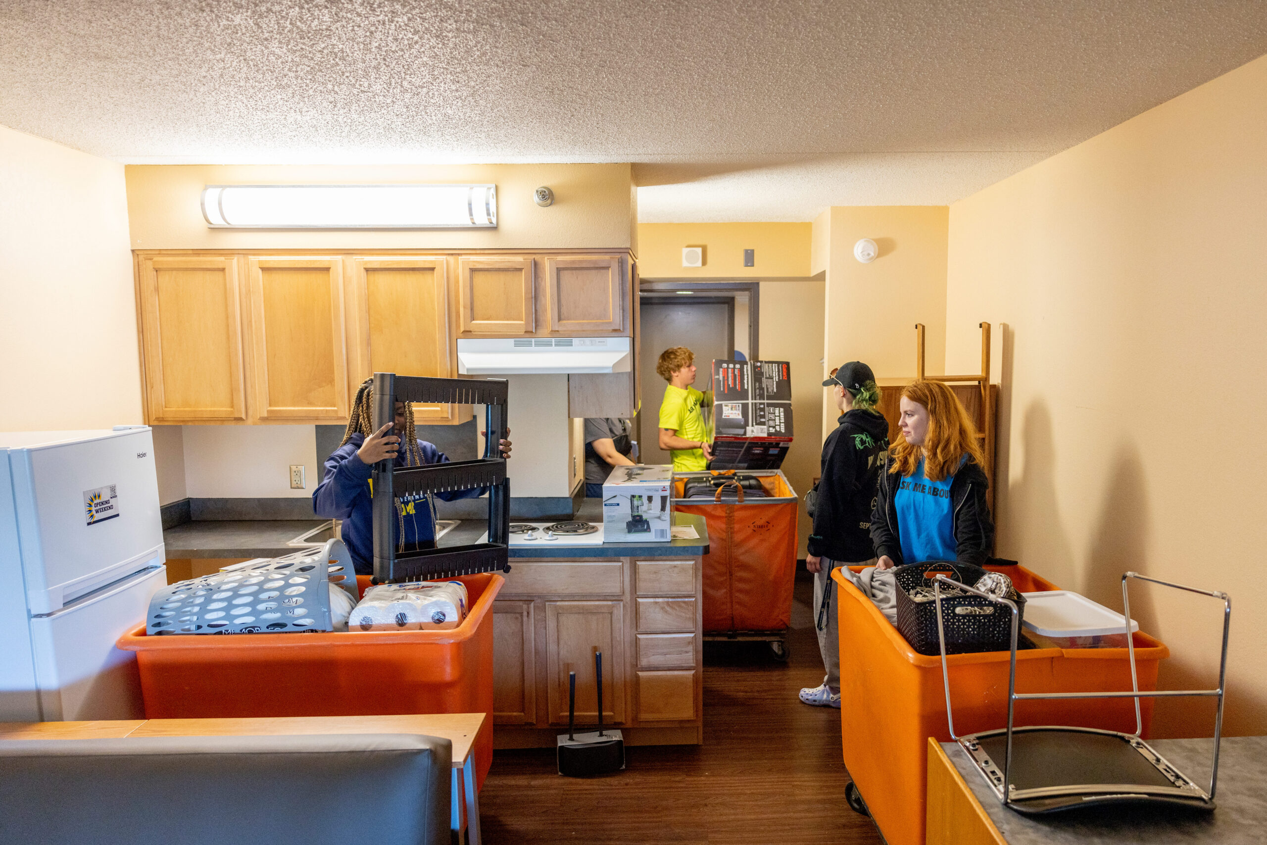 A group of four people is in a kitchen area, moving items into orange carts. One person holds a black shelf, while another carries a box. The kitchen has wooden cabinets and a white refrigerator.