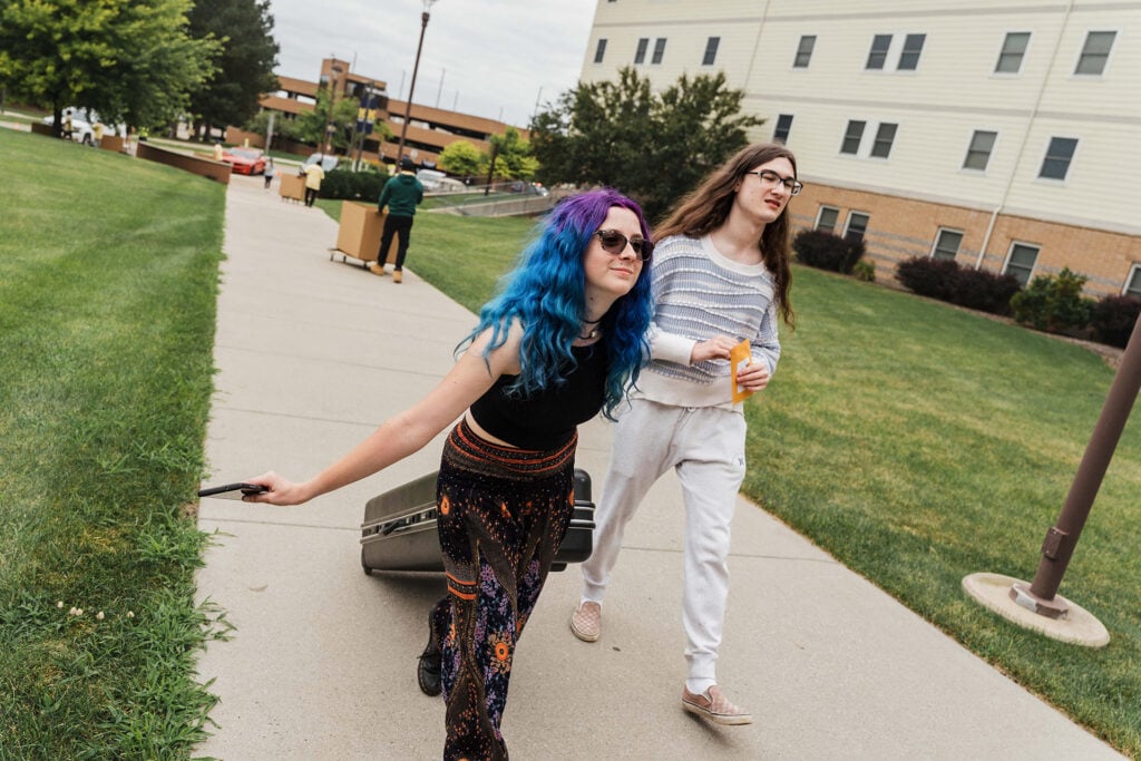 Two people walk along a sidewalk, one pulling a suitcase and the other holding a yellow envelope.