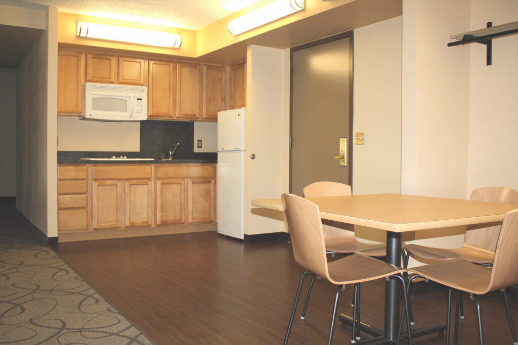 Small kitchen area with wooden cabinets, white microwave and refrigerator, and a dining table with four wooden chairs.