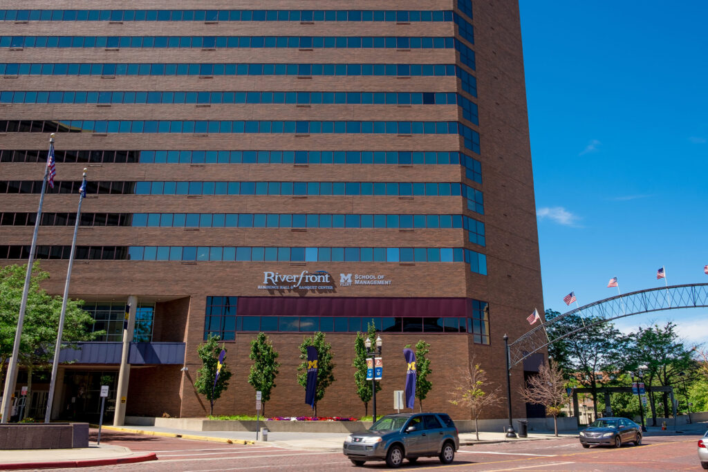 A view of the Riverfront Residence Hall and Banquet Center, featuring large windows, trees, and flags, with the School of Management sign visible.