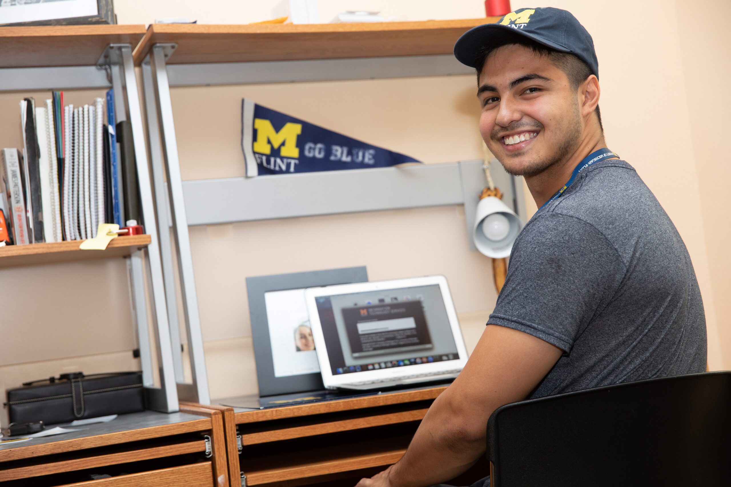 A person sitting at a desk with a laptop, surrounded by books and a pennant that says "M FLINT GO BLUE."
