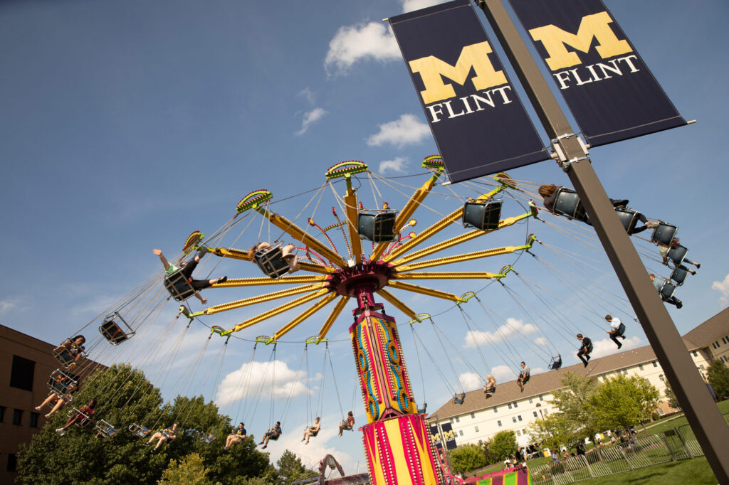 People riding a colorful swing ride at an outdoor event with University of Michigan-Flint banners visible.