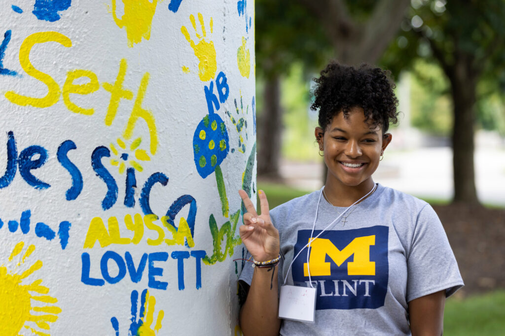 Person wearing a gray University of Michigan Flint t-shirt making a peace sign next to a white wall with colorful handprints and names painted on it.