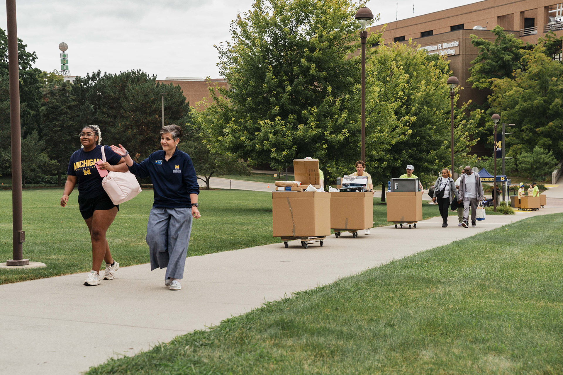 Two people walk on a path, one carrying a bag, while others push carts loaded with boxes. Green trees and a building are in the background.