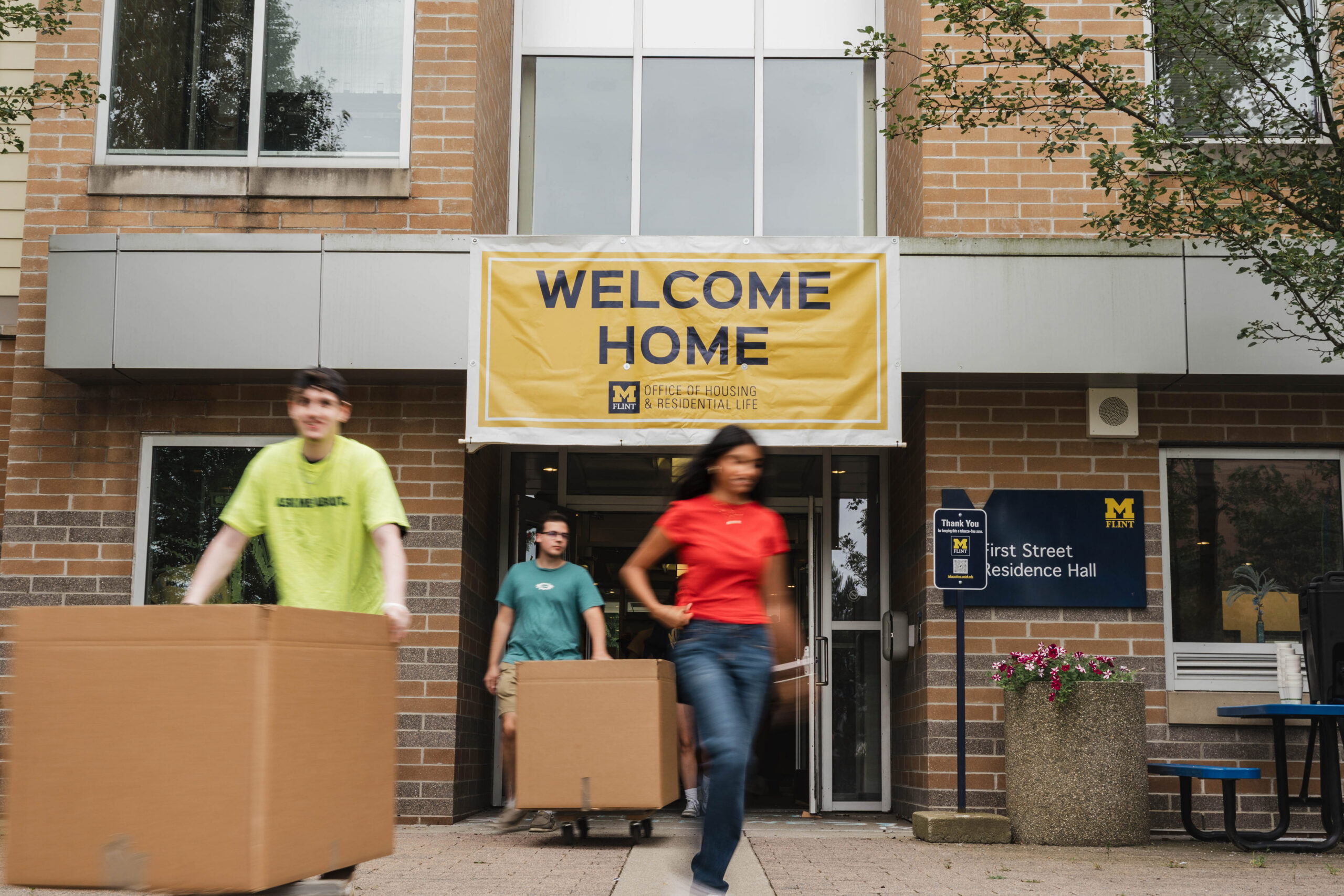 A banner reading "WELCOME HOME" hangs above the entrance of a residence hall, as three individuals carry boxes outside.