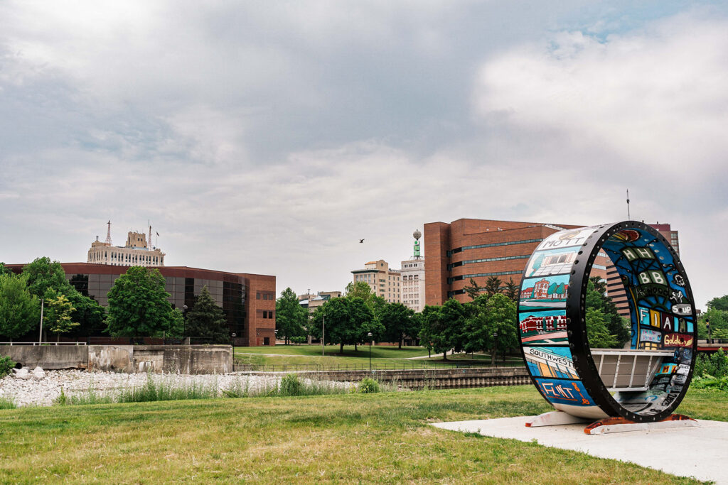 A colorful circular sculpture with various painted designs stands in a park, with buildings and trees in the background.