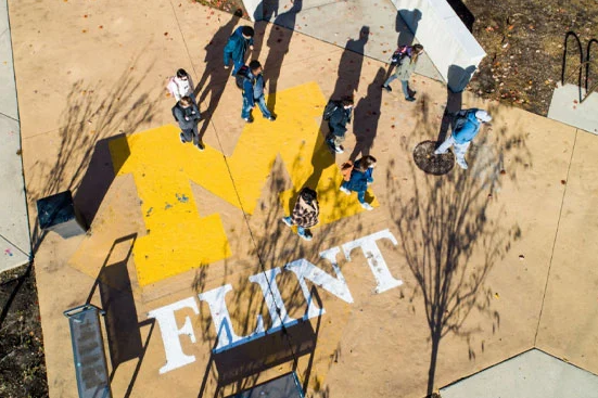 Aerial view of a group of people gathered around a large yellow "M" and the word "FLINT" painted on the ground.