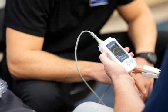 A person holds a medical device displaying readings while another person's arm is visible nearby.