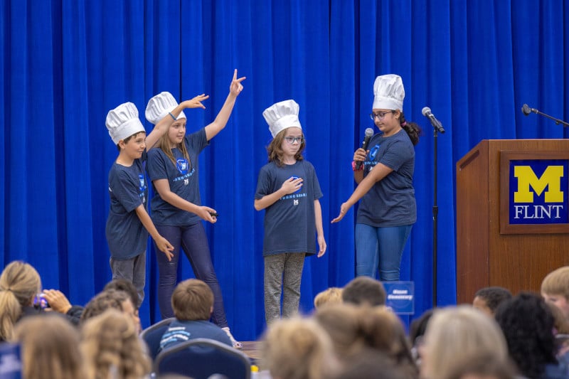 Four children wearing chef hats perform on stage in front of an audience, with a blue curtain and a podium in the background.