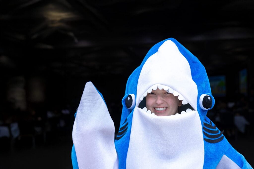 A person in a blue shark costume smiles widely, with a blurred background of a gathering.