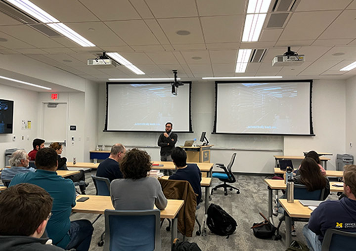 A person stands in front of a classroom with two large screens, addressing a group of seated students.
