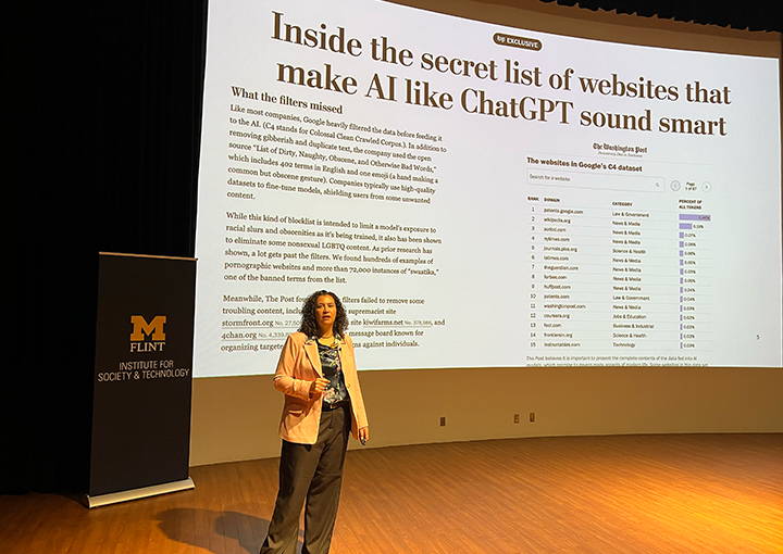 A person stands on stage in front of a large screen displaying an article titled "Inside the secret list of websites that make AI like ChatGPT sound smart."
