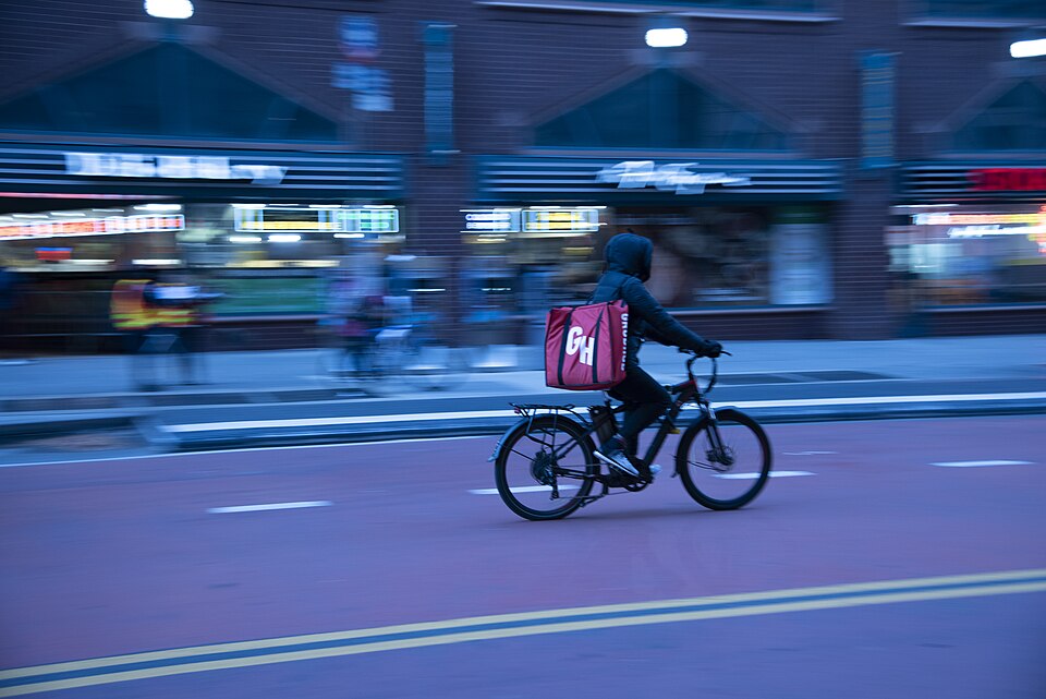 A person rides a bicycle on a city street, carrying a large delivery bag on their back, with blurred pedestrians in the background.