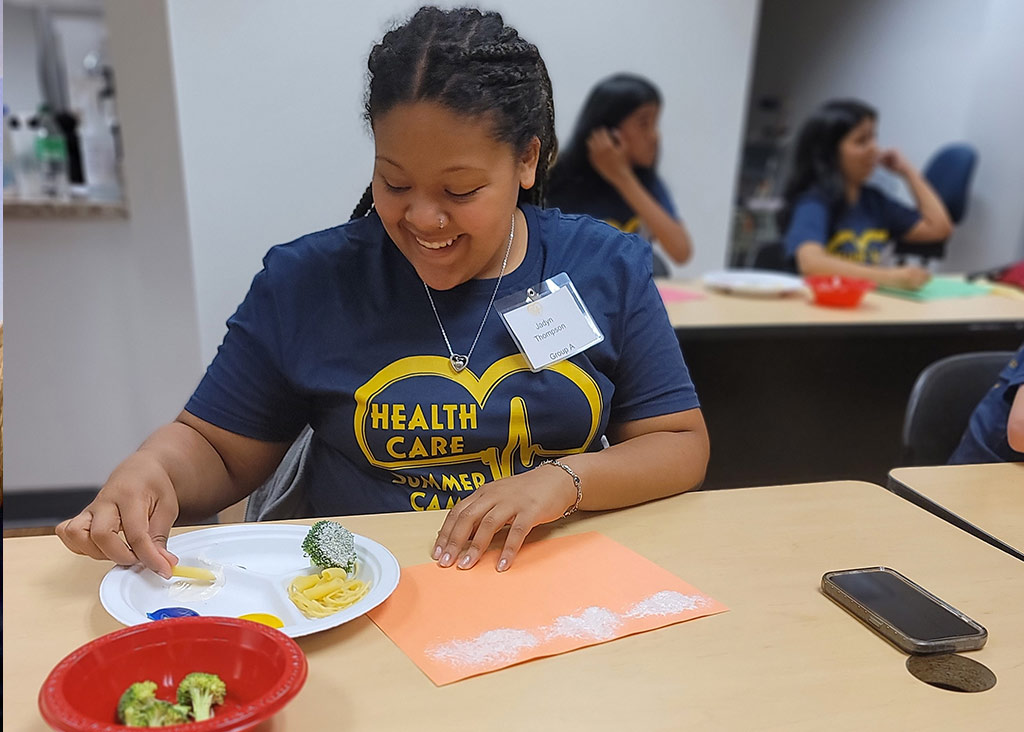 A young woman in a "Health Care Summer Camp" shirt smiles while doing an activity with food and paper.