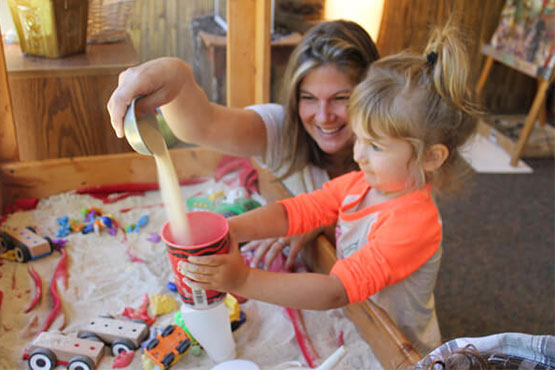 A young child and an adult woman engage in a sensory play activity, pouring sand into a cup in a bright, wooden play area.