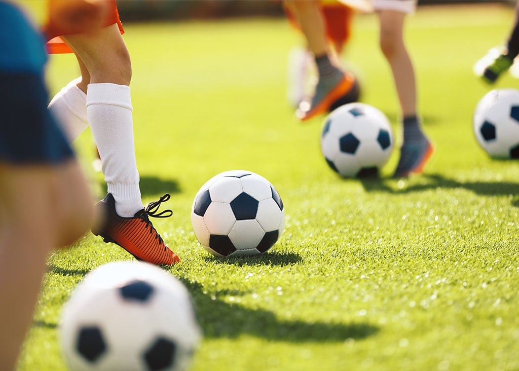 Children playing soccer on a grassy field with multiple soccer balls visible.