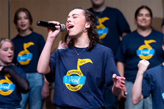 A young woman sings passionately into a microphone on stage, surrounded by a choir wearing matching "Summer Vocal Academy" shirts.