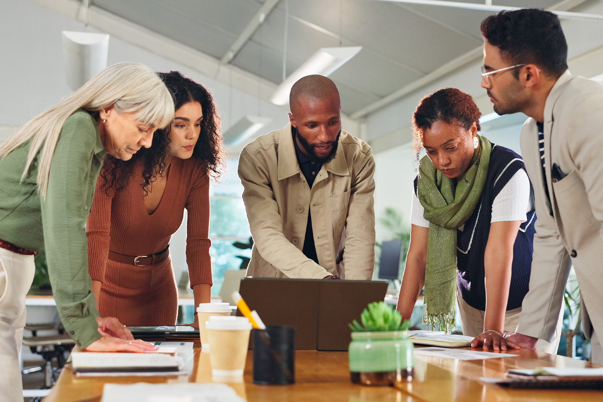A group of five people is gathered around a laptop on a table, discussing and collaborating on a project.