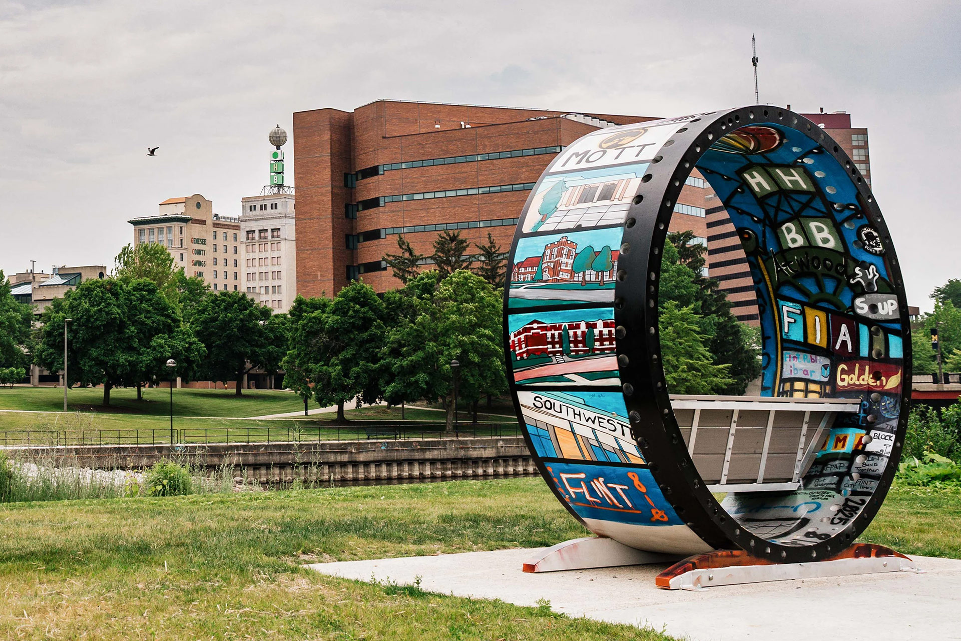 A colorful, artistic bench shaped like a large wheel, with various designs and text, set in a park with buildings in the background.