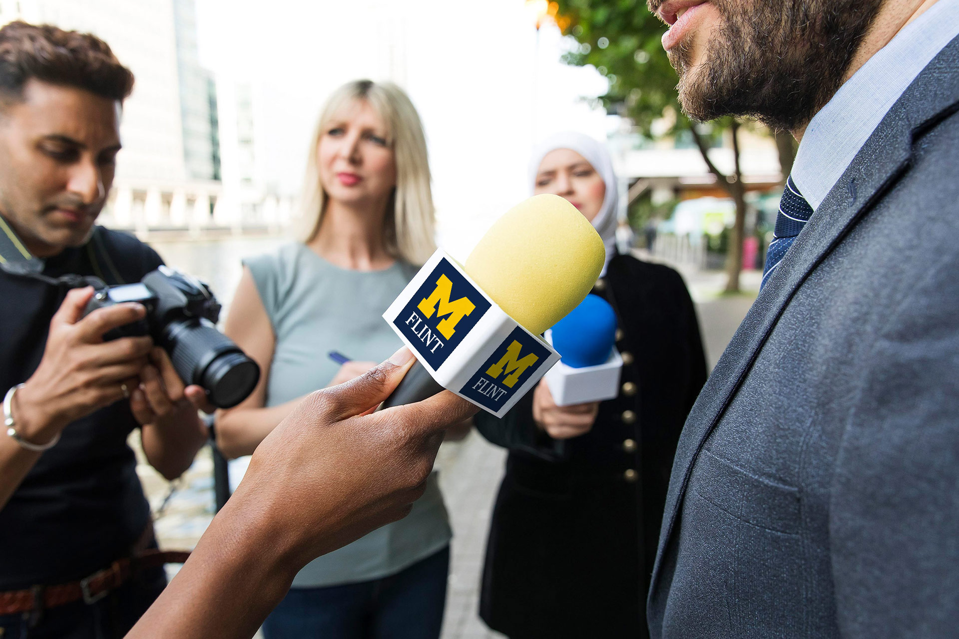 A person holds a microphone with "M FLINT" while another person is being interviewed outdoors, with a camera nearby.