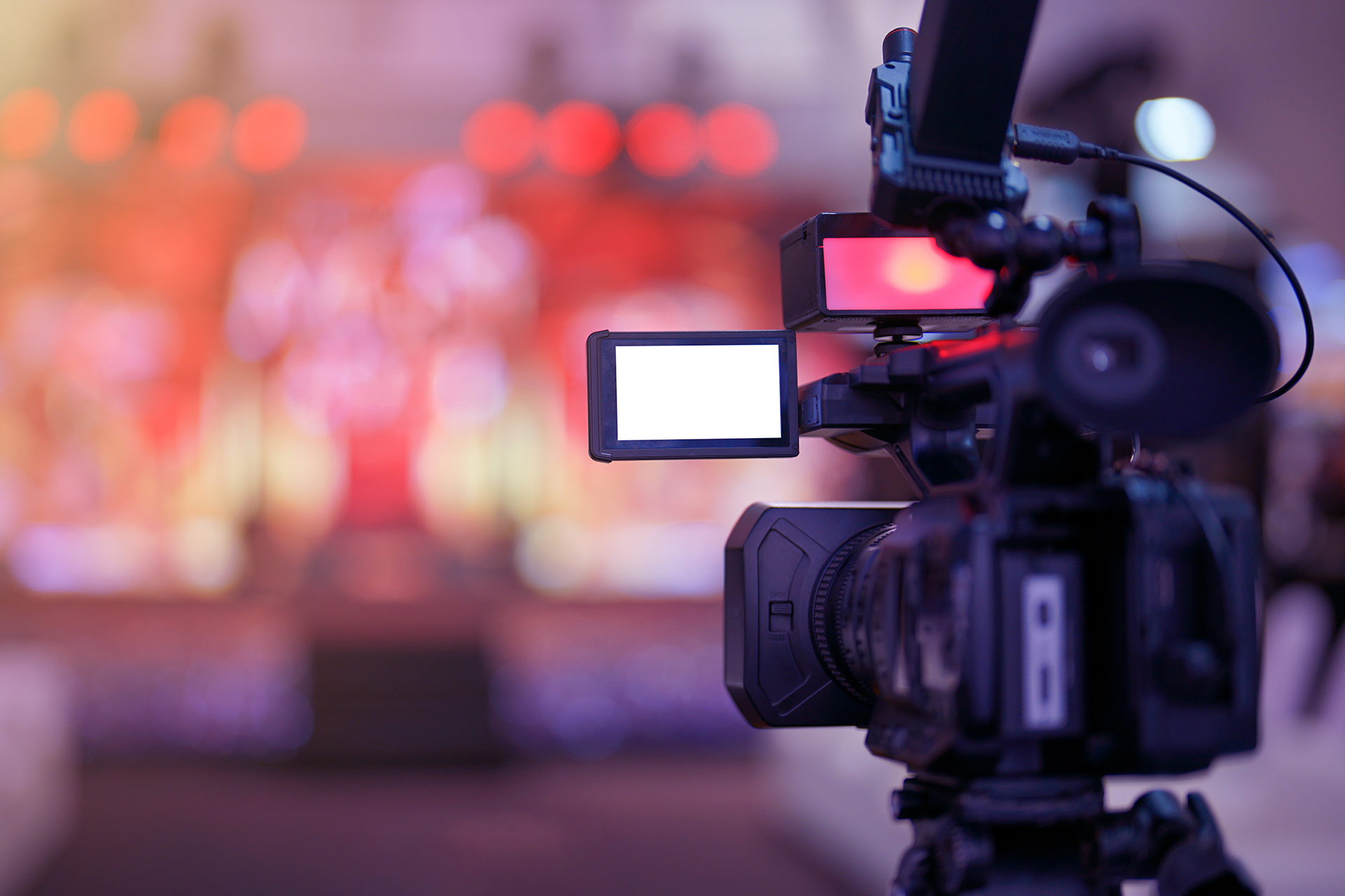 A video camera with a display screen is positioned in the foreground, with a blurred stage and lights in the background.