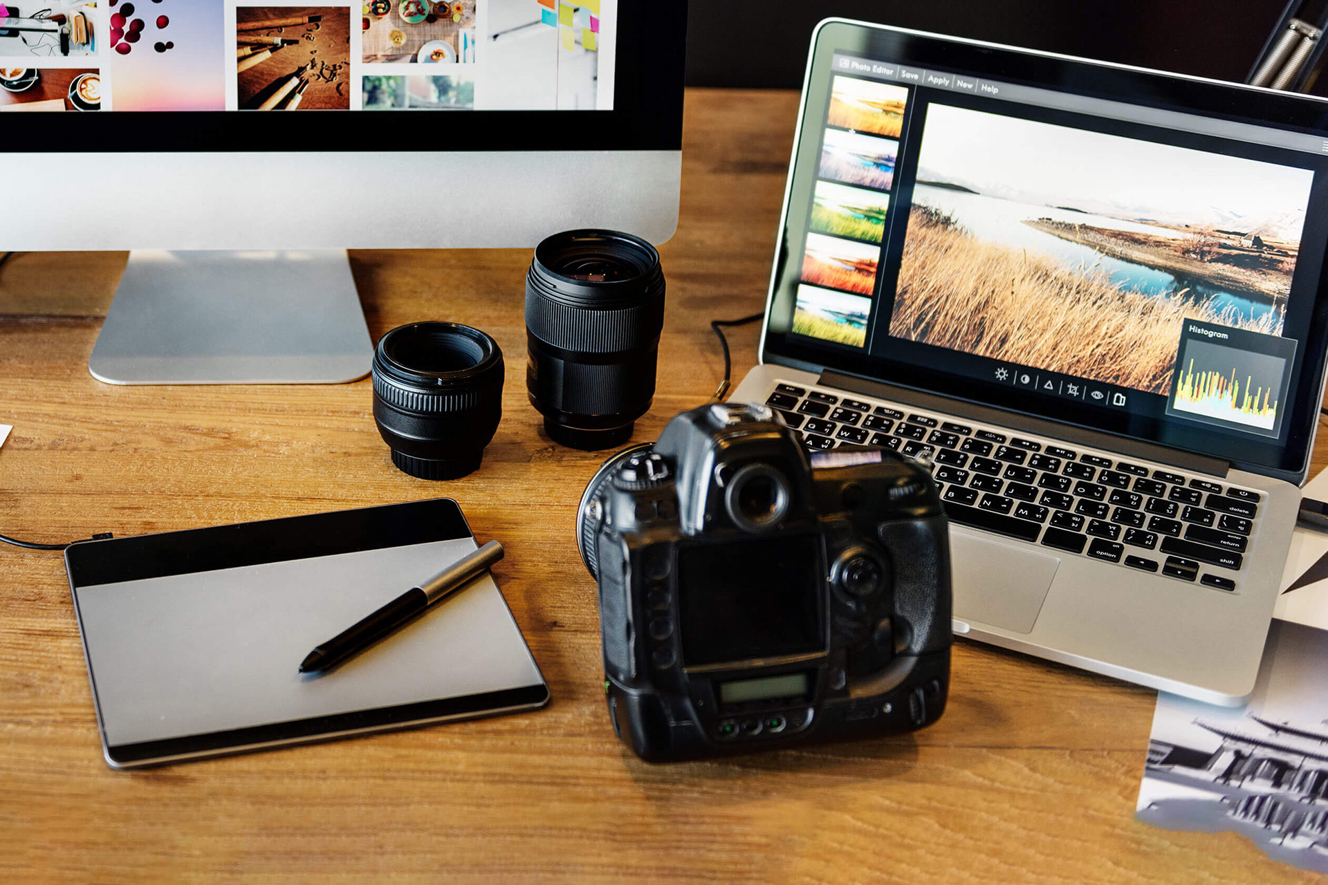 A workspace featuring a laptop displaying photo editing software, a camera, two camera lenses, a graphics tablet, and a pen.