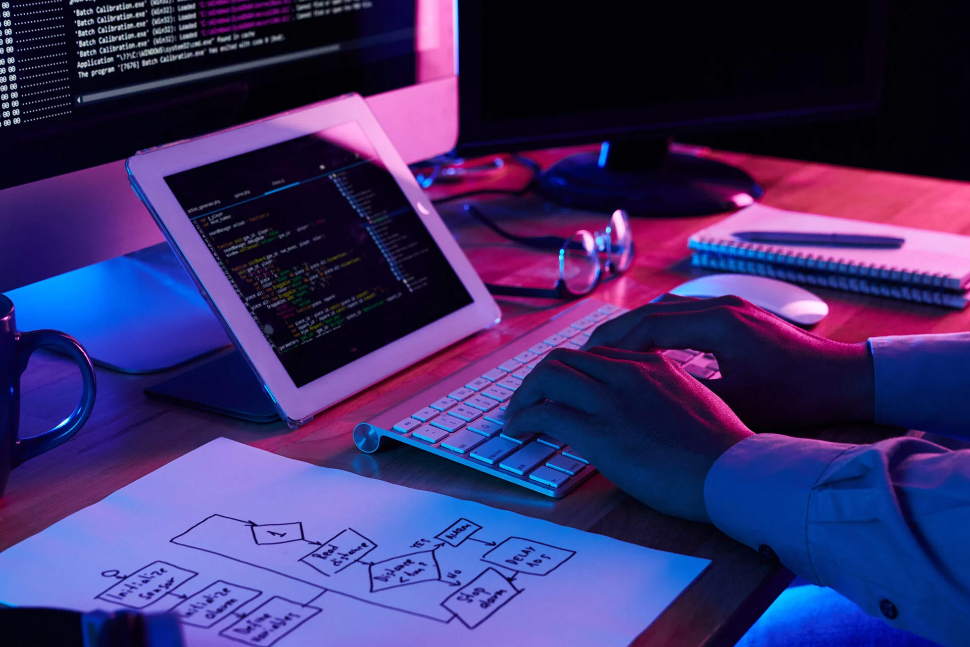 A person types on a keyboard in a dimly lit workspace with a tablet displaying code, a flowchart on paper, and a mug nearby.