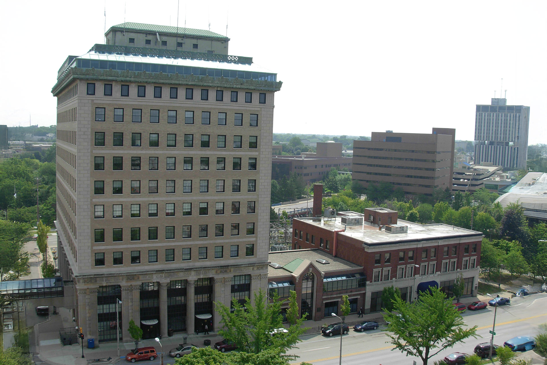 A view of a cityscape featuring a tall building with a flat roof, surrounded by greenery and other buildings in the background.