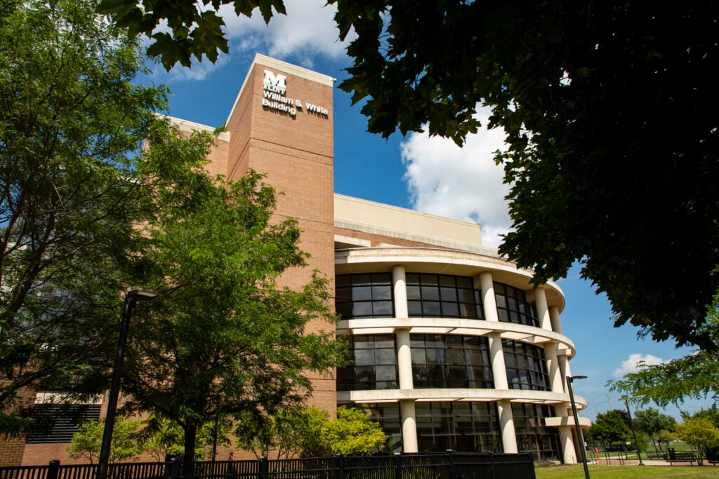 The image shows the exterior of the William S. White Building, surrounded by trees and a clear blue sky.