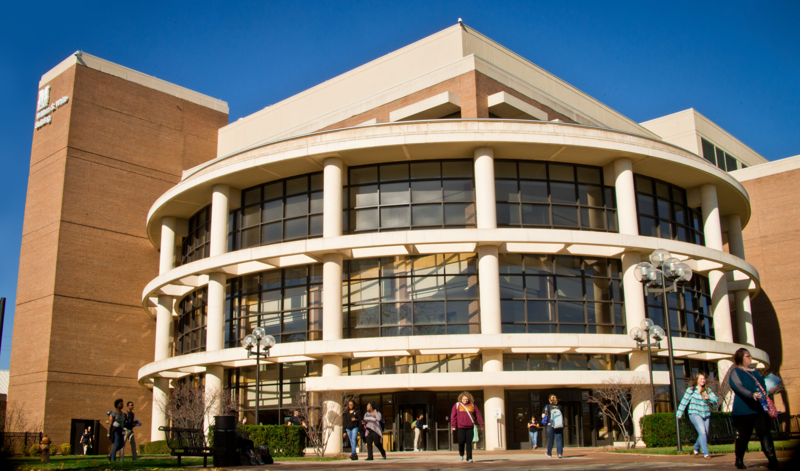 A modern building with large windows and a circular design, with several people walking in front on a sunny day.