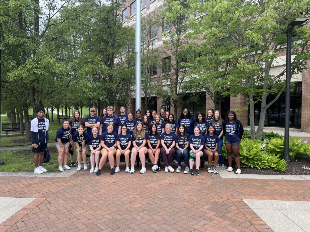 A group of people poses outdoors in matching shirts that read "FUTURE SUMMER CAMP," surrounded by trees and a building.