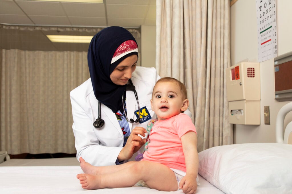 A healthcare professional examines a seated baby in a medical setting, with medical equipment visible in the background.