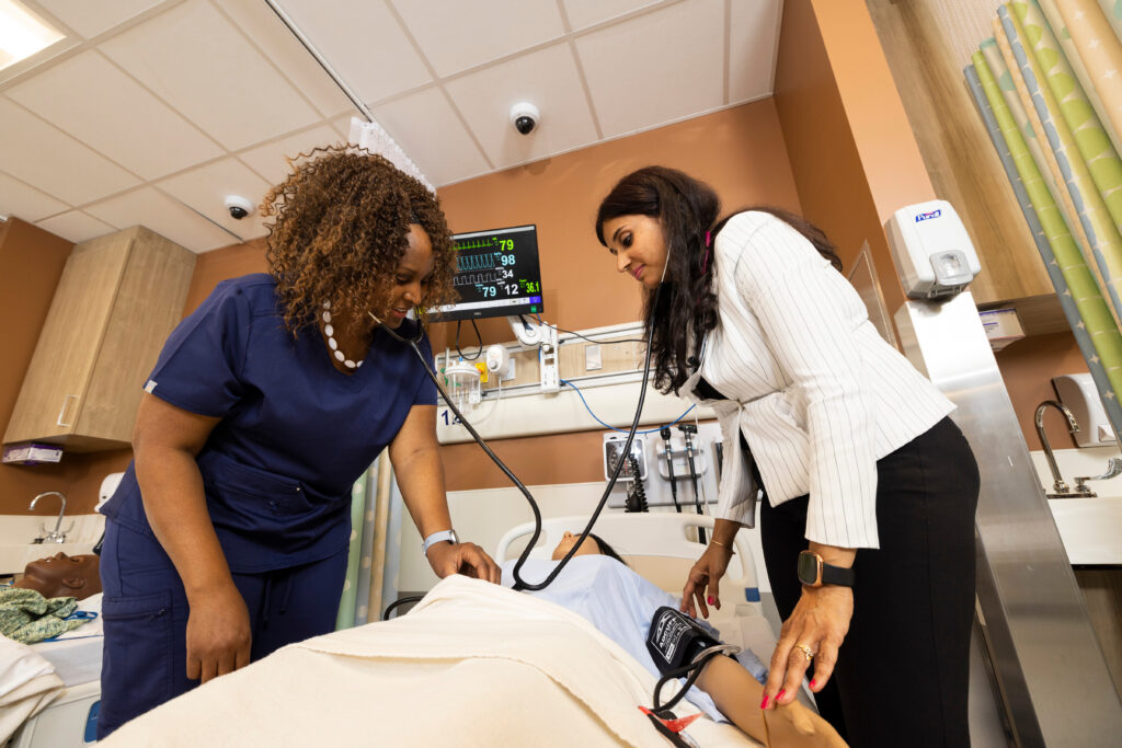 Two healthcare professionals are checking a patient's vital signs in a hospital room, with medical equipment visible in the background.