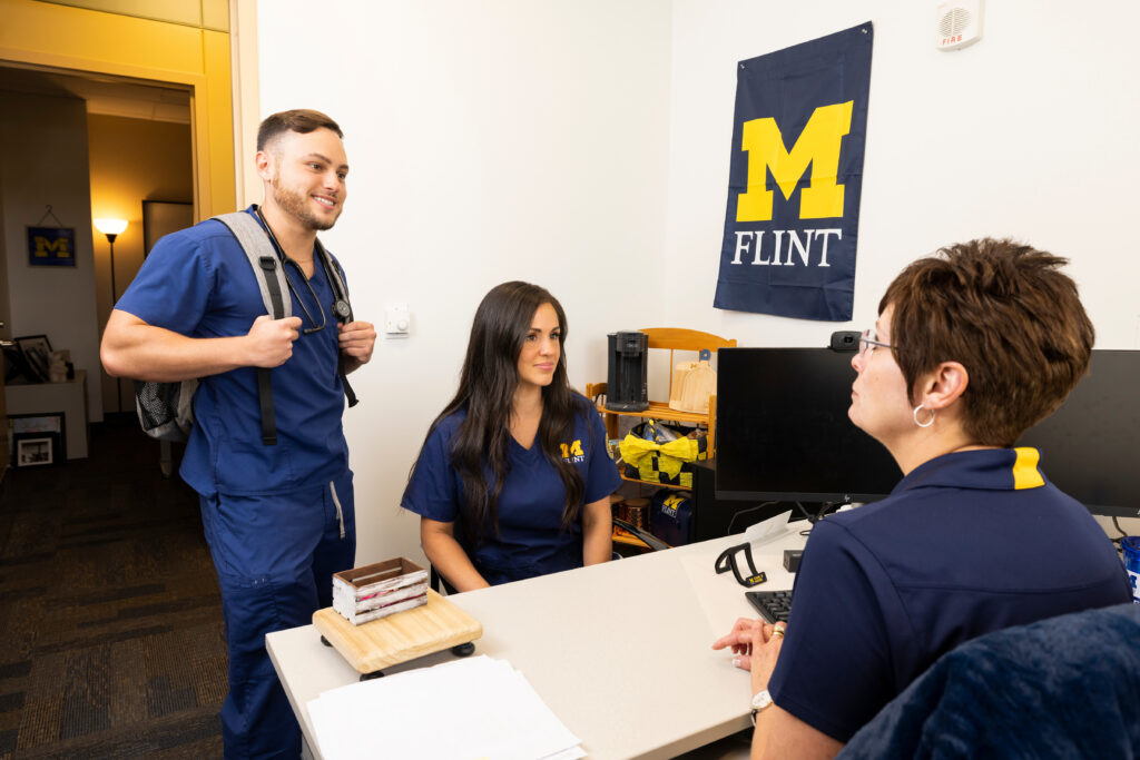 A person in scrubs with a backpack talks to two others at a desk in an office, with a university flag on the wall.