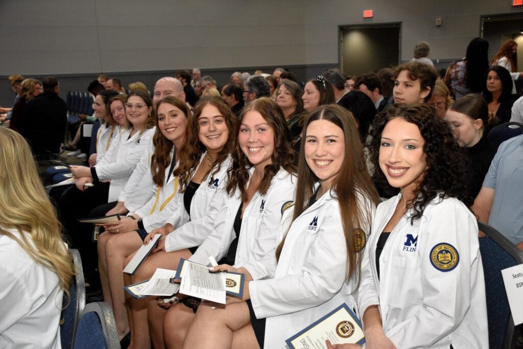 A group of seven people in white coats smile while holding certificates, seated in an audience during a ceremony.