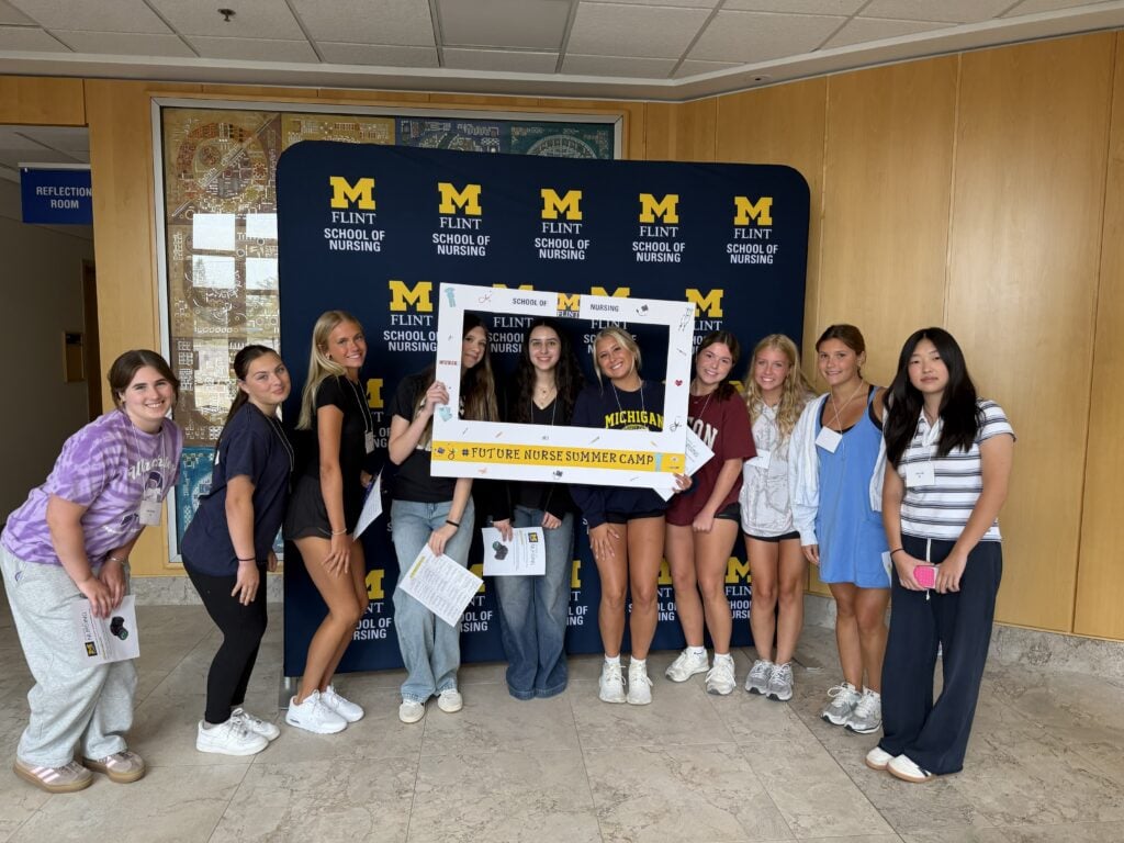 A group of people poses together with a frame that reads "#FUTURE NURSE SUMMER CAMP" in front of a backdrop for the Flint School of Nursing.