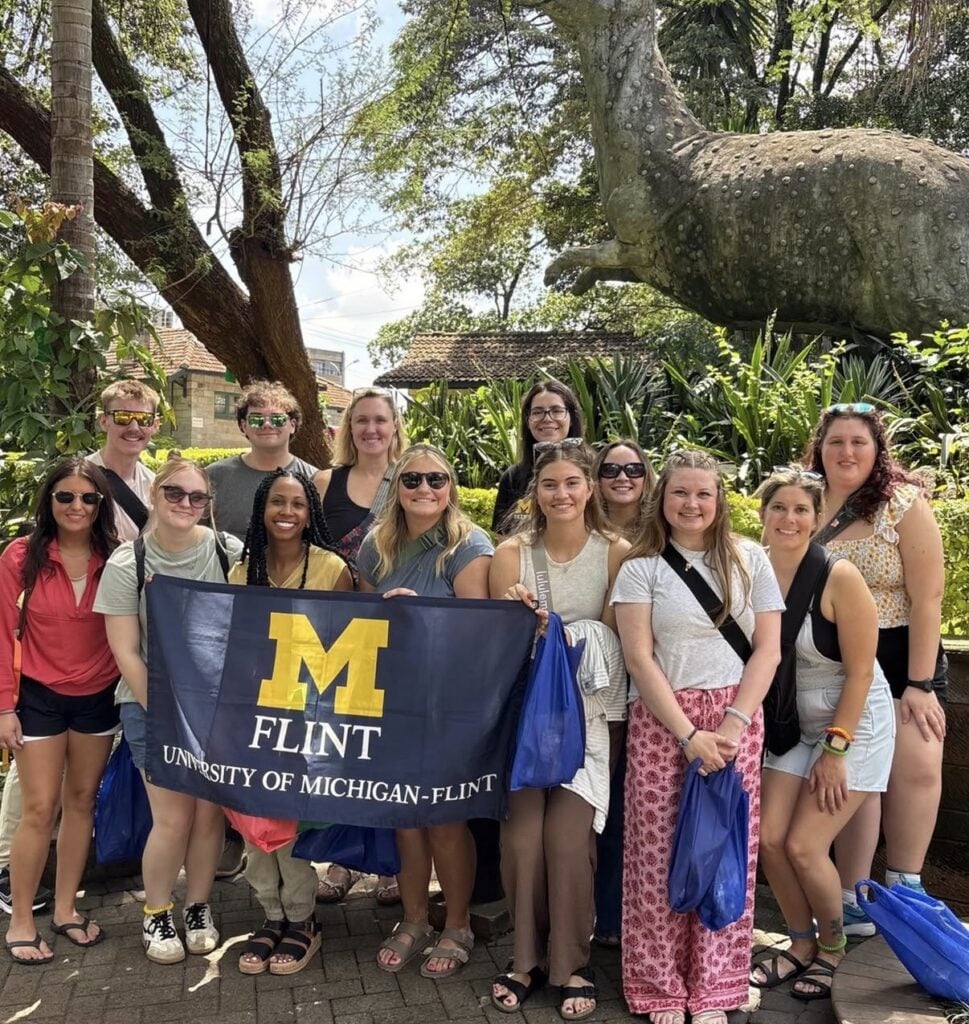 A group of people poses outdoors holding a University of Michigan-Flint banner, with a dinosaur statue in the background.