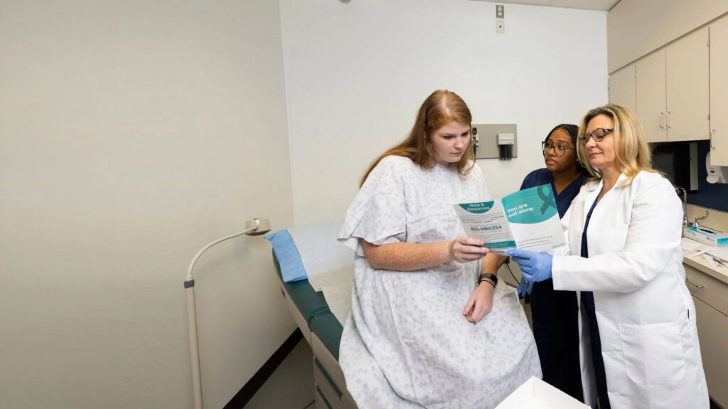 A person in a hospital gown sits on an exam table, holding a brochure while two healthcare professionals discuss it.